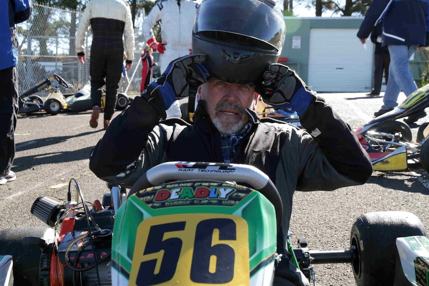 A man sitting on a go-kart puts on a helmet.