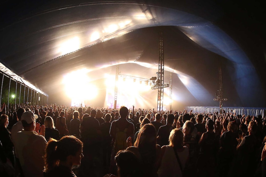 A crowd watches a performer on a brightly lit stage at a music festival.
