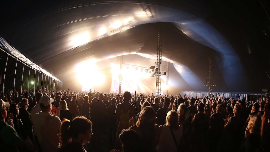 A crowd watches a performer on a brightly lit stage at a music festival.