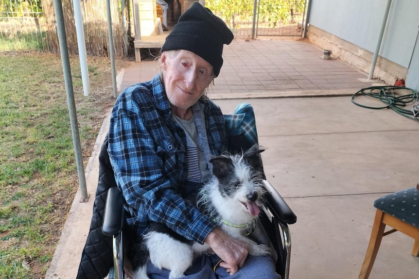 A man wearing a blue flannel shirt smiles at the camera, with his favourite dog on his lap. The dog is black and white.
