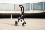 A white man with long hair in a pony tail and black t-shirt and shorts taking a step with the help of a walker