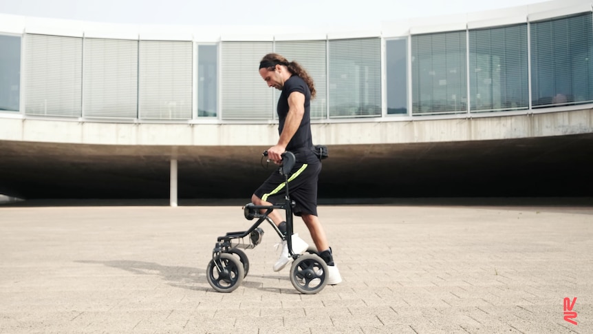 A white man with long hair in a pony tail and black t-shirt and shorts taking a step with the help of a walker