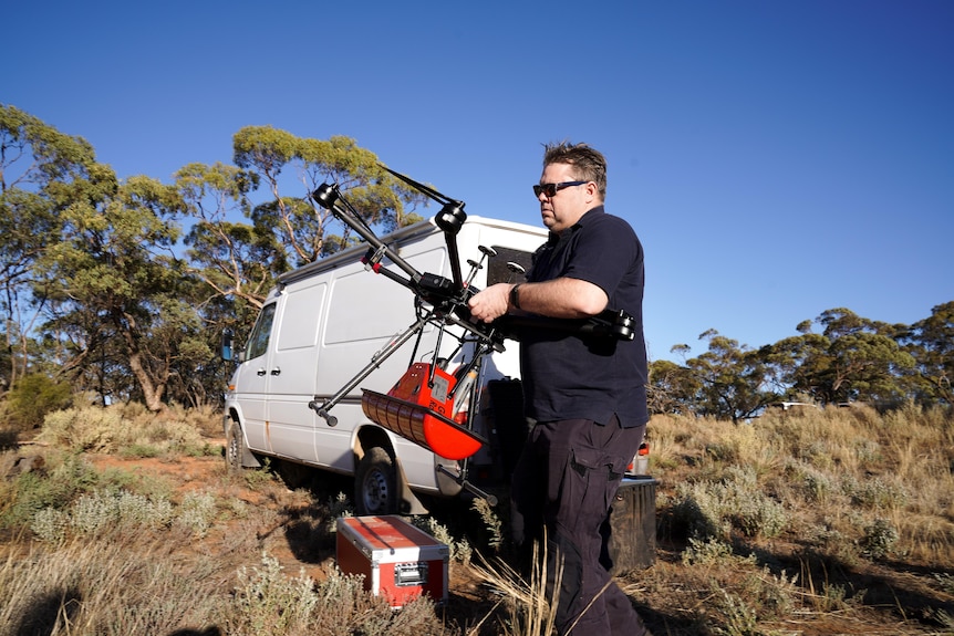 A man holding a drone in front of a van and scrubland