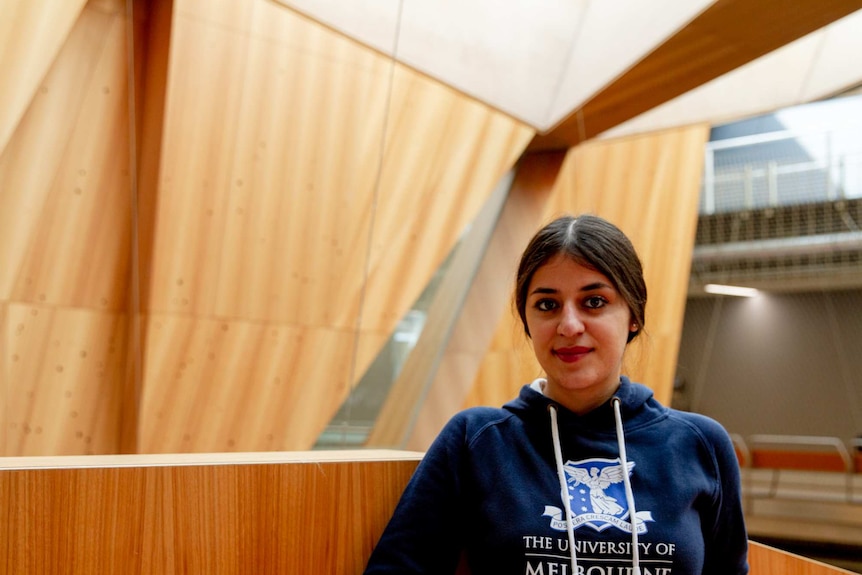 You view a woman looking directly at the camera in a brown top, in front of a brown brick wall.