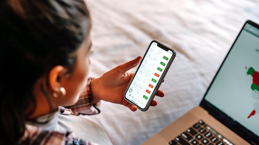 A woman looks at a stock market chart on a laptop while sitting on a bed.