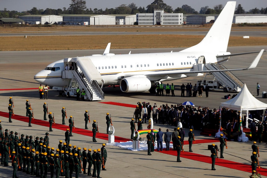 On an airfield lies a white Zimbabwean plane as rows of soldiers flank the tarmac which carries the coffin of Robert Mugabe.