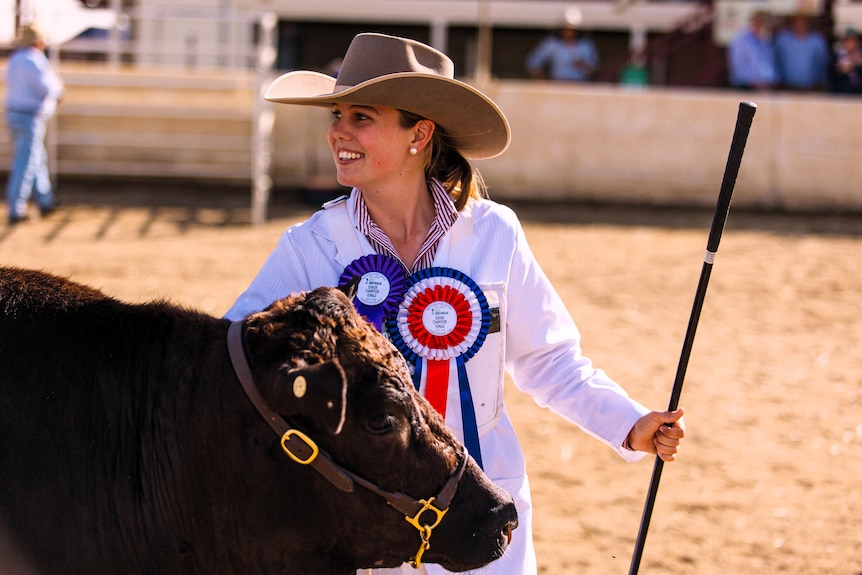 A young woman smiling and wearing a winning ribbon as she holds onto stud cattle.