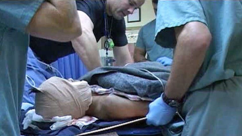 TV still of head bandaged patient surrounded by staff at Role Three Hospital in Afghanistan