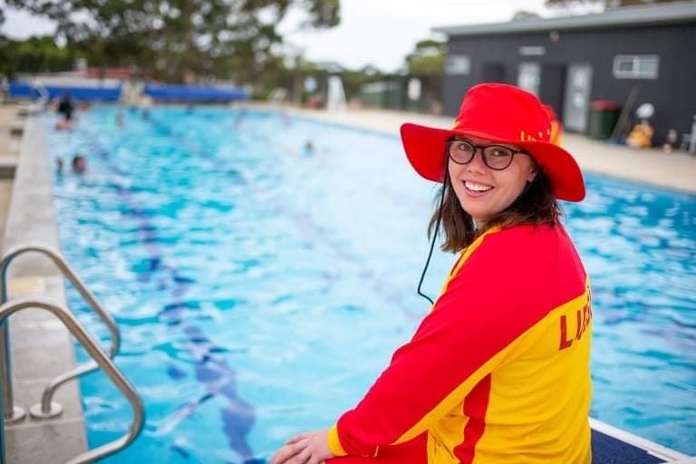 A white woman with glasses smiling, wearing a red and yellow lifeguard uniform in front of a pool.