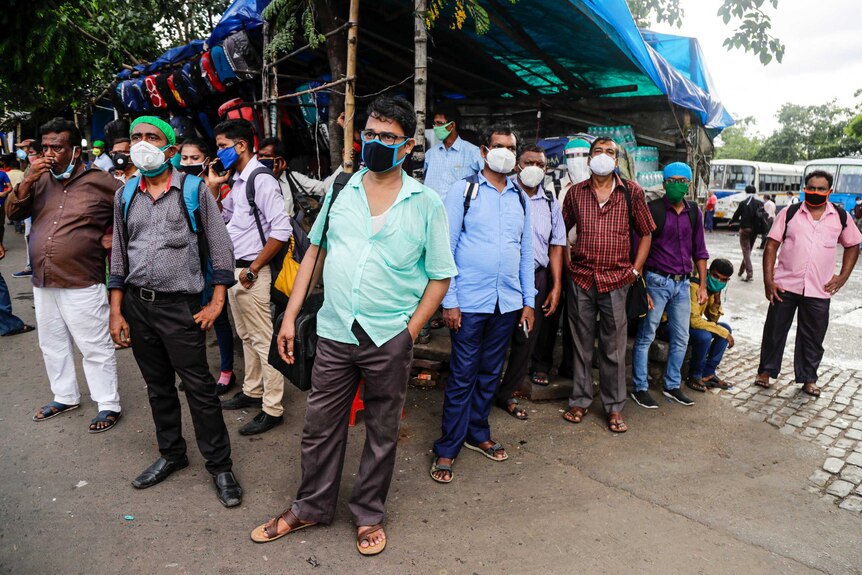 People wearing face masks look in the same direction as they wait for a bus on the side of a road.