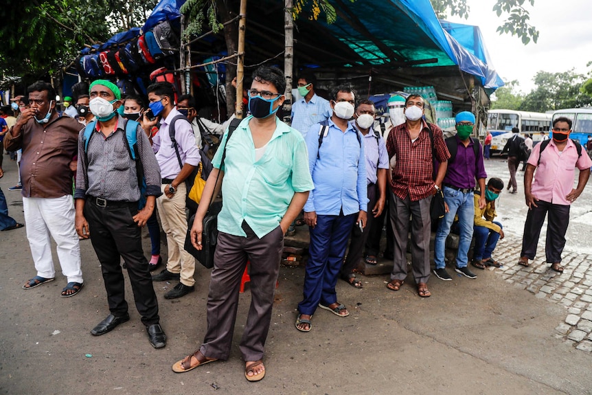 People wearing face masks look in the same direction as they wait for a bus on the side of a road.