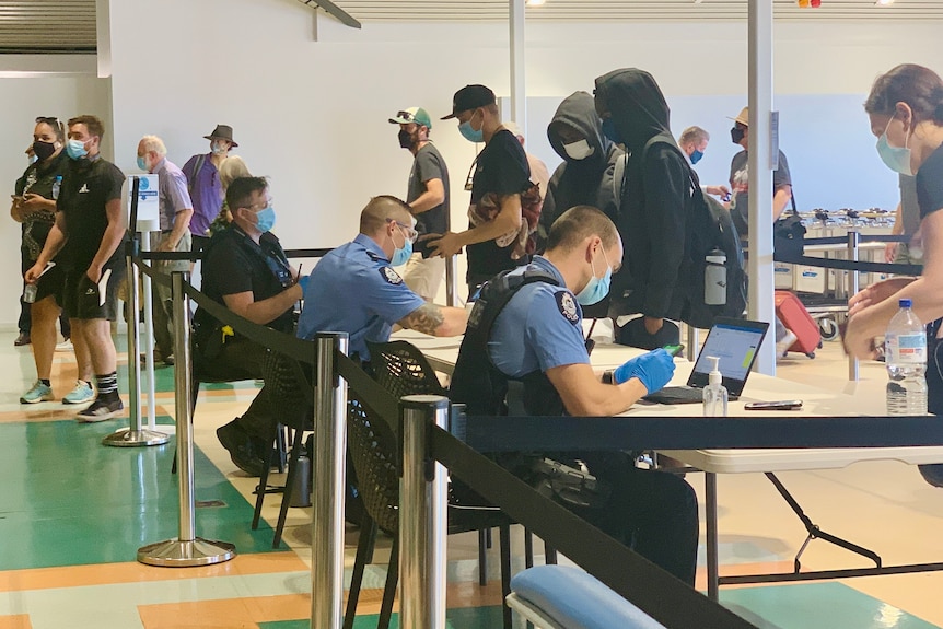 Two police officers wearing face masks sit in front of queueing aeroplane passengers in a crowded airport terminal