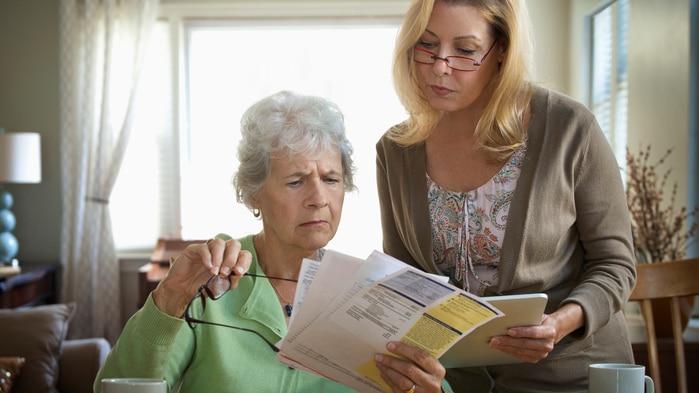 An older woman looks worriedly at some bills while a younger woman, presumably her daughter, looks over her shoulder.