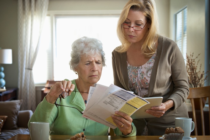 An older woman looks worriedly at some bills while a younger woman, presumably her daughter, looks over her shoulder.
