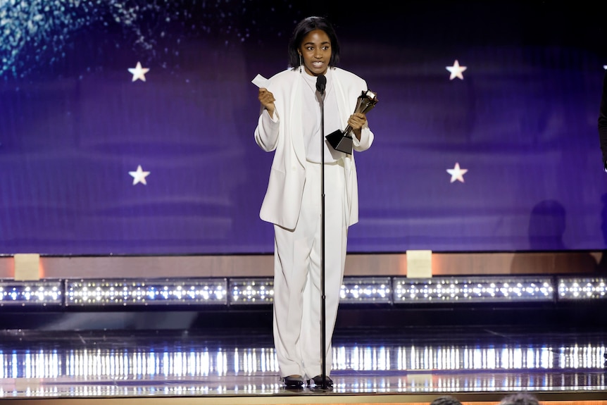 Ayo, dressed in an oversized white suit, holds up her speech as she accepts her award on stage.