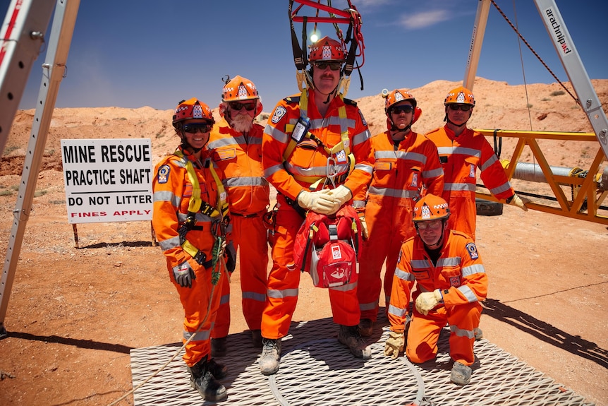 A group in orange protective clothing stand together in front of a mine rescue practice shaft