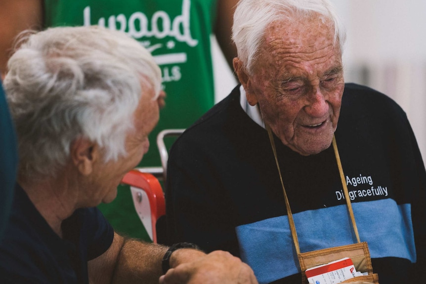 An elderly man sitting in a wheelchair talks to another man with white hair in the foreground.