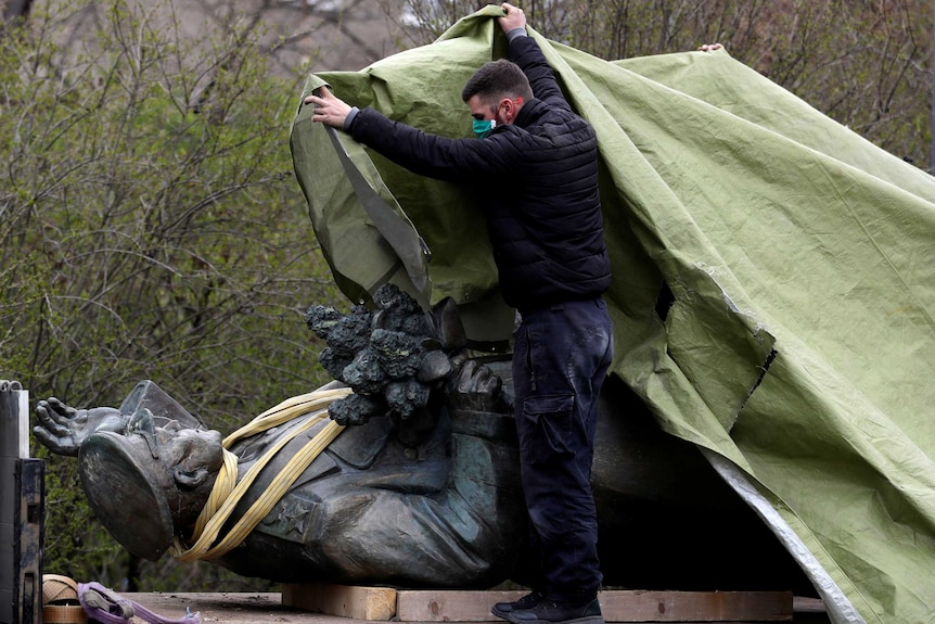 A man in a face mask covers a dark bronze statue of a man in military uniform with a tarp. The statue is on its back.