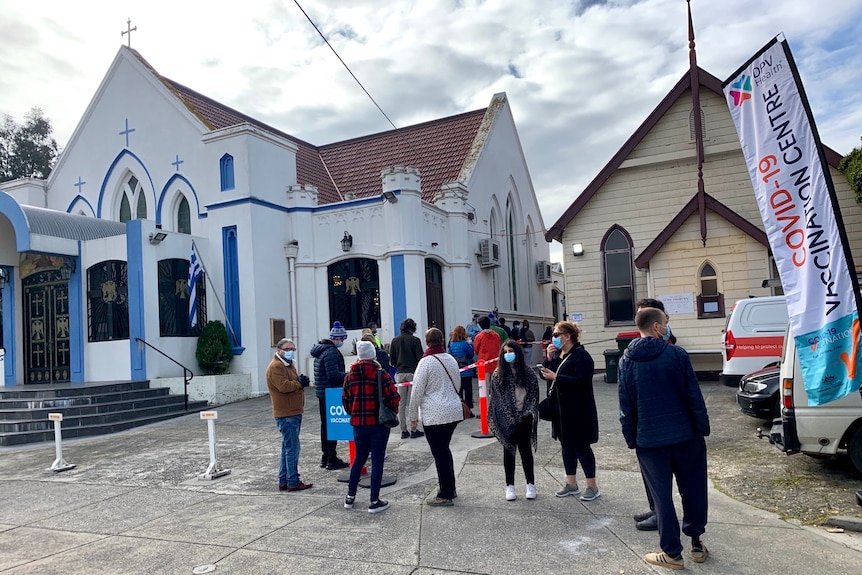 A distanced line of people with masks queue outside a church beside a sign reading 'COVID-19 vaccinations'.