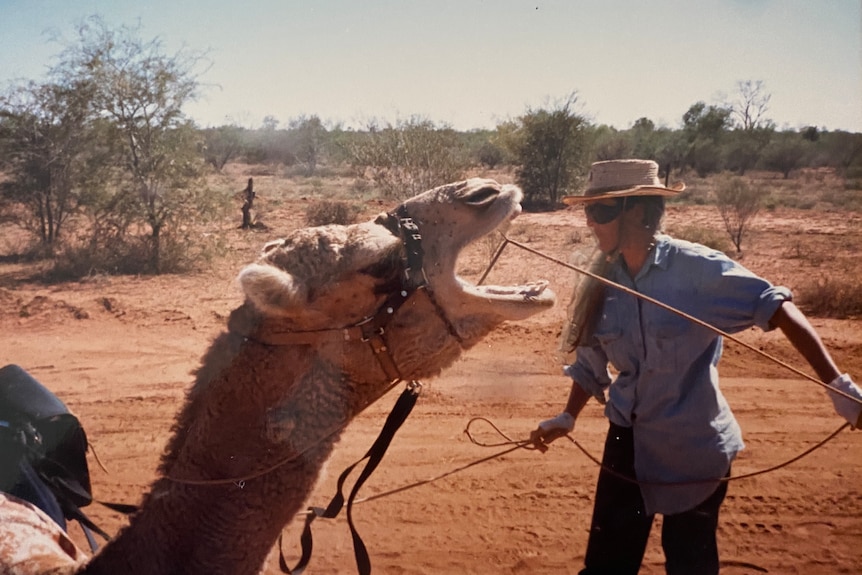 Photo of Jill Colwell surrounded by red dirt pulling the reins of a camel with its mouth open.