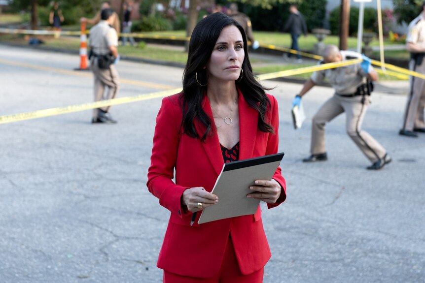 A dark-haired woman in her 50s wearing a red suit holds a notepad as she stands at the yellow taped perimeter of a crime scene