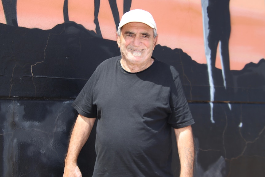 A smiling man wearing a black shirt and white hat standing in front of a mural