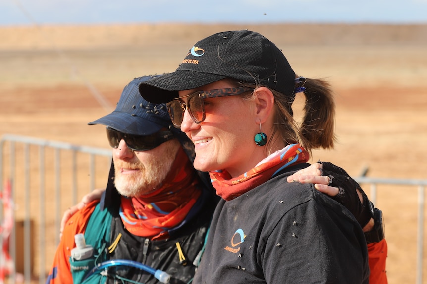 A man in running gear and a woman in a black hat smile while covered in flies.