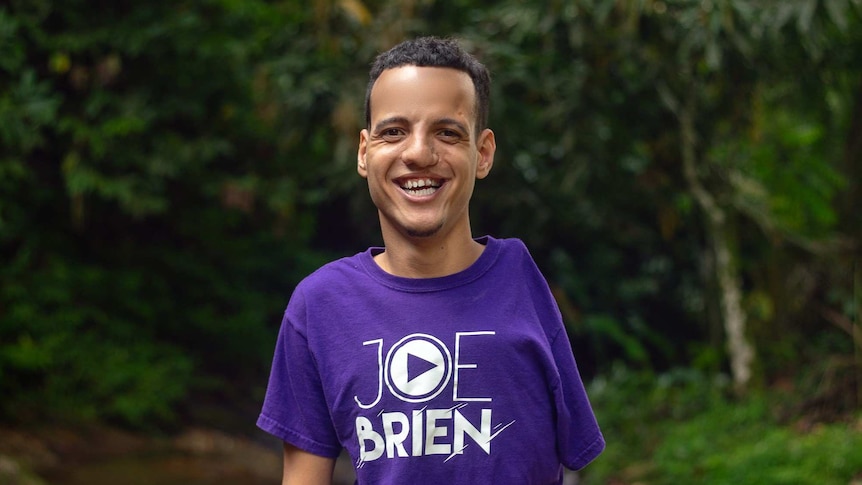 Joe Brien wears a purple tee and stands with trees in the background. He has one arm, is 3 feet tall, and wears grey shoes.