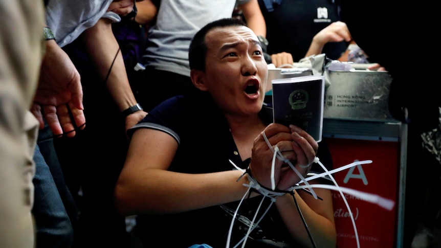 A man with zip ties around his wrists holds his passport and shouts as he's surrounded by people