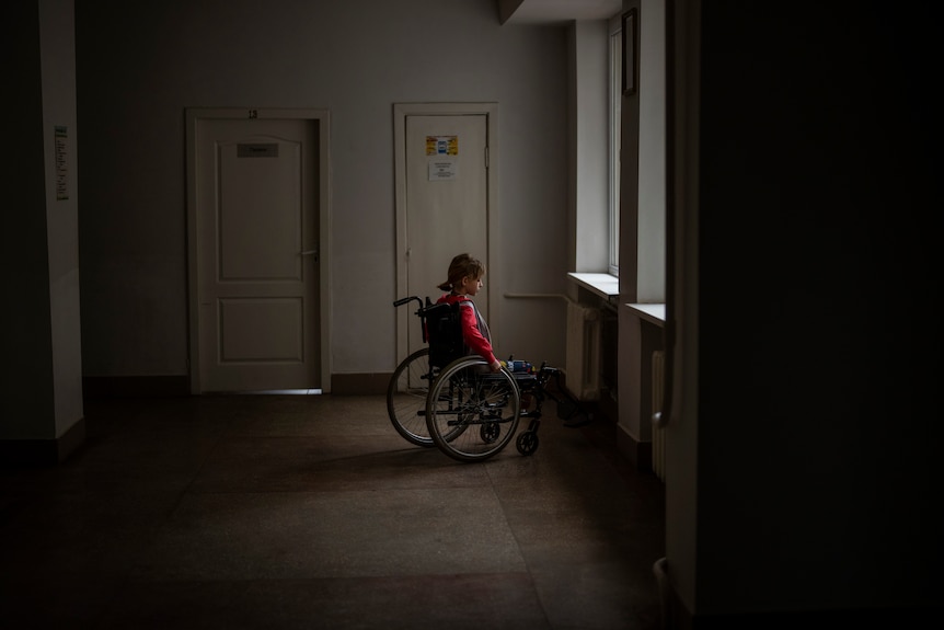 In a darkened corridor, a girl sits in a wheelchair chair looking out of one of two windows 
