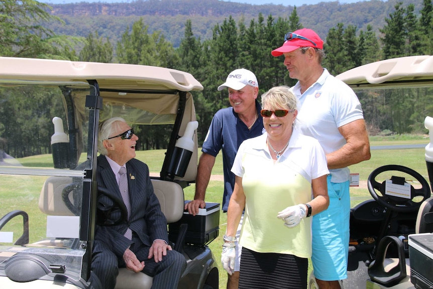 Three golfers stand to the side of a golf cart talking to a centenarian