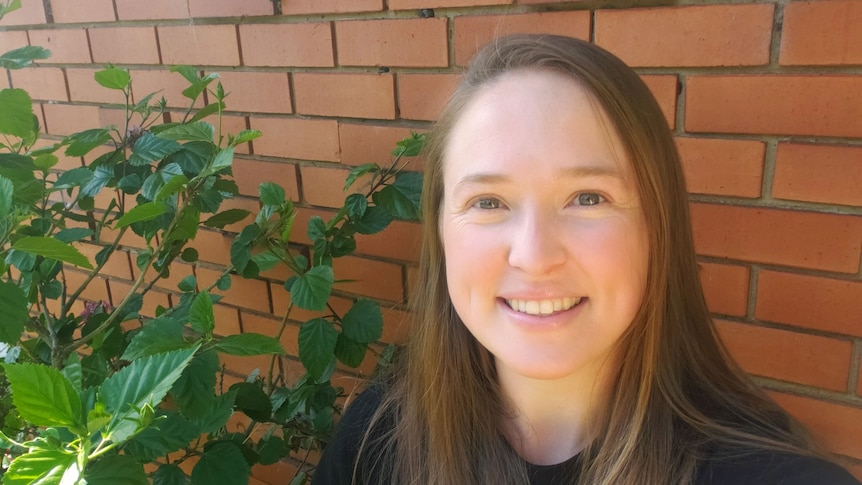 Ellen stands in front of a wall at her home while smiling.