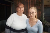 A woman and her adult daughter stand side by side, with an arm around each other, looking into the camera.