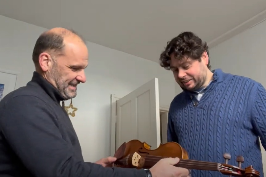 Two men stand together, both looking at a violin being held in one man's hands.