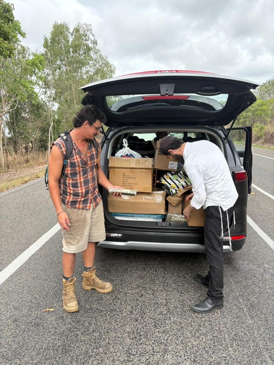 Dos hombres se acercaron al maletero de un coche lleno de bolsas y cajas en una carretera.