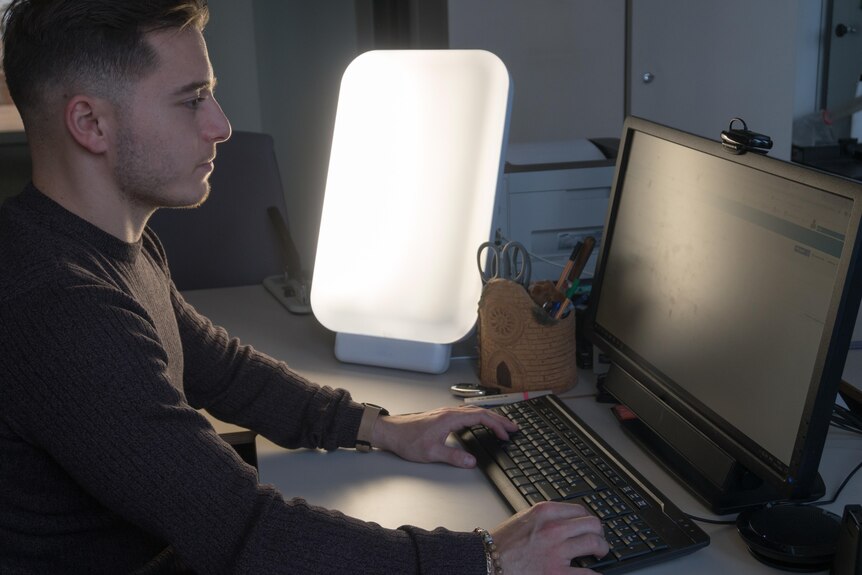 A man sits at a desk working at a computer with a light box to the side of him