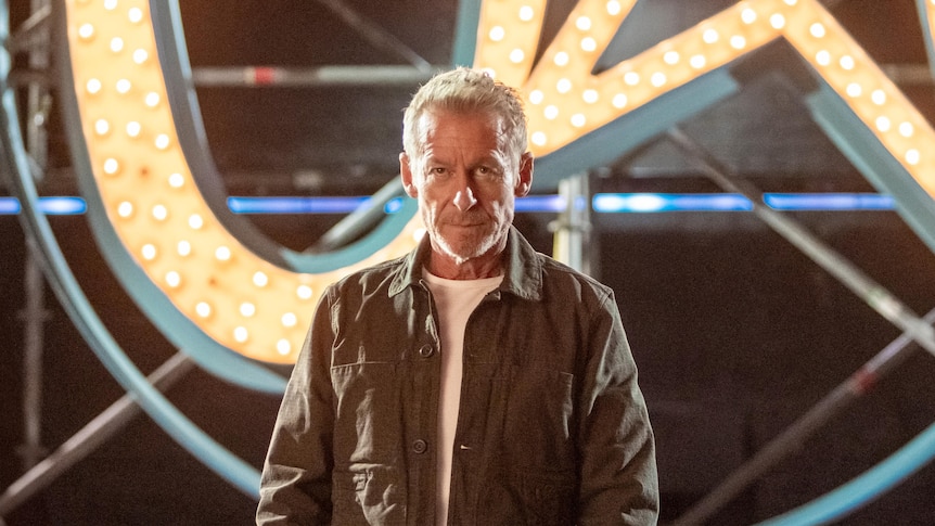 Richard Roxburgh stands in front of scaffolding and a lit-up neon U Star logo of a U and a star shape in a circle.