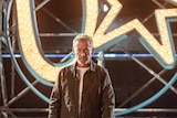 Richard Roxburgh stands in front of scaffolding and a lit-up neon U Star logo of a U and a star shape in a circle.