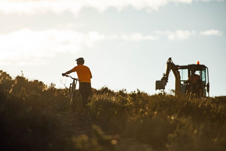 A man pushing a bike across heath covered ground to access a small working excavator.