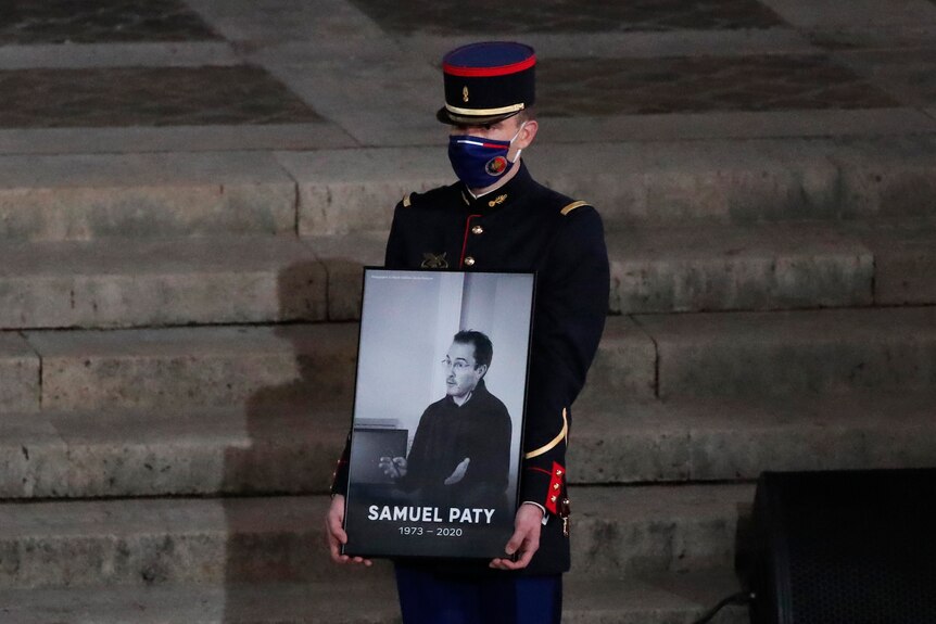 A man in uniform holds up a picture of a man, underneath it reads "Samuel Paty 1973-2020"