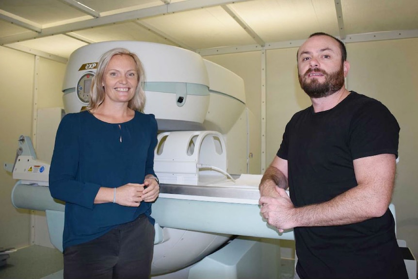 A woman and man stand and smile in front of a large bulky white MRI machine