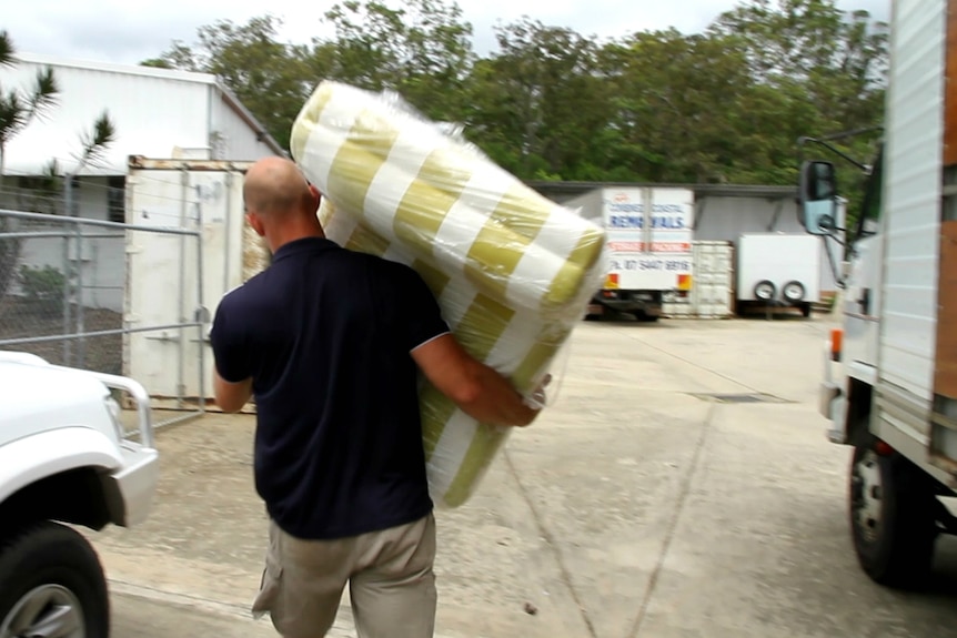 A man walking away carrying a striped lounge chair wrapped in plastic.