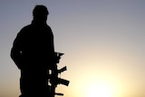 Soldier stands in desert field holding service weapon with sunrise behind him