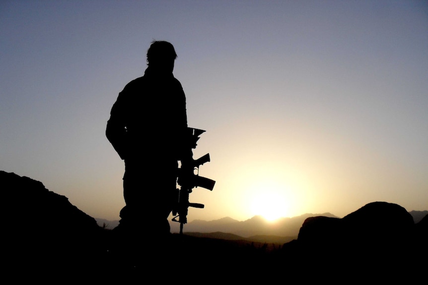 Soldier stands in desert field holding service weapon with sunrise behind him