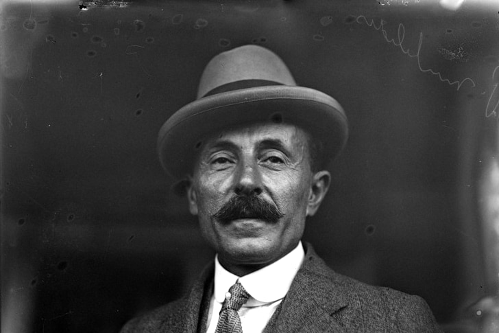 A black and white head and shoulders portrait-style image of a moustached man in a suit and hat.