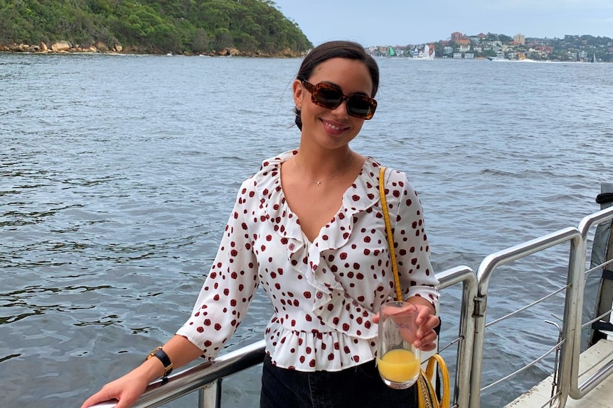 A woman wearing sunglasses and holding a glass of orange juice stands on a pontoon on Sydney Harbour.