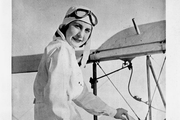 A black and white magazine front cover of a female pilot sitting in a plane with goggles on.