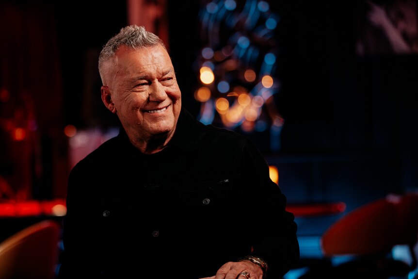 A landscape image of Jimmy Barnes wearing a black top. The background is out of focus but there is red, orange and blue light