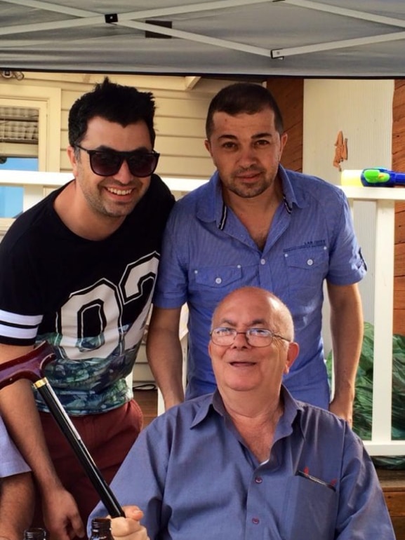 Apostolis Barbousas holds a cane and sits smiling with two of his sons standing behind him.