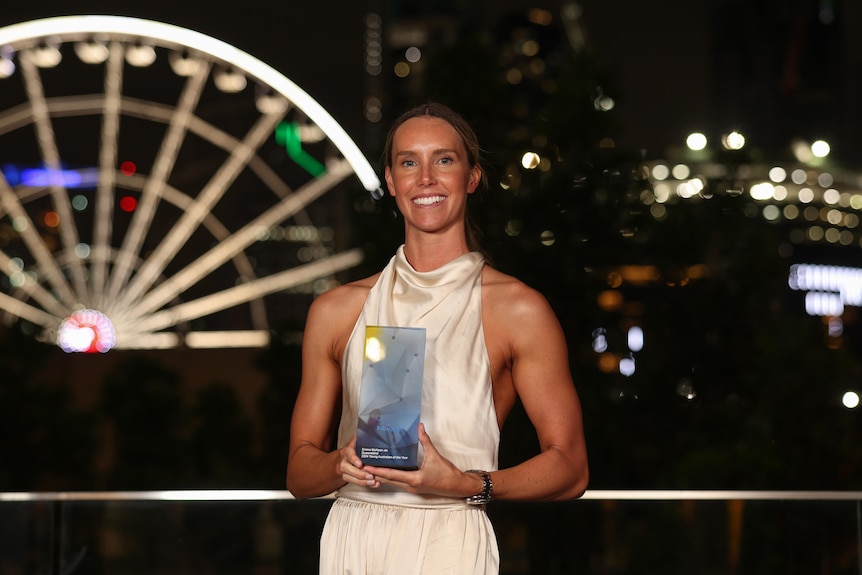 A woman in a white jumpsuit holds a glass award with a lit up ferris wheel in the background.